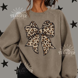 May include: A taupe-colored crewneck sweatshirt featuring a large, centered graphic of a bow with a brown and black leopard print pattern. The sweatshirt has long sleeves and a ribbed neckline, with a watermark logo visible on the fabric.