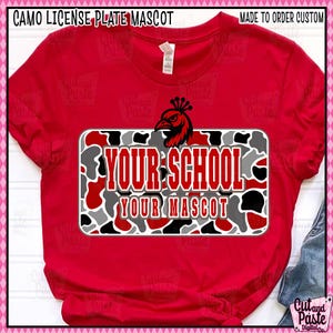 May include: Red t-shirt with a license plate design that reads "YOUR SCHOOL YOUR MASCOT". The license plate has a camouflage pattern in red, grey, and black. A black and red bird graphic is above the text.
