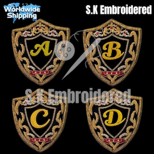 Könnte beinhalten: Vier gestickte Schildaufnäher, jeder mit einem anderen Buchstaben: A, B, C und D. Die Schilde haben einen schwarzen Hintergrund mit goldenen und roten Details. Die Buchstaben sind in einer gelben, stilisierten Schriftart. Der Text "S.K Embroidered" ist sichtbar.