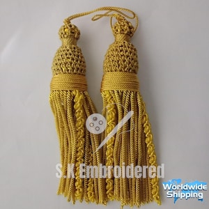Peut inclure: Deux glands dorés avec des dessus tissés complexes et de longues mèches fluides. Les glands sont reliés par un cordon doré. Le texte "S.K Embroidered" est visible en bas de l'image.