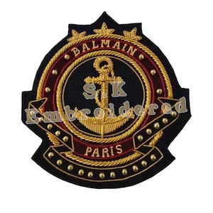 Op de afbeelding: Een zwarte geborduurde patch met een gouden anker, touw en kroon. De tekst "BALMAIN PARIS" is in goud geborduurd rondom het anker. De patch is versierd met gouden studs.