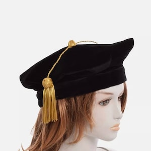 May include: Black velvet academic cap with a gold tassel and cord. The cap is worn on a mannequin with brown hair.