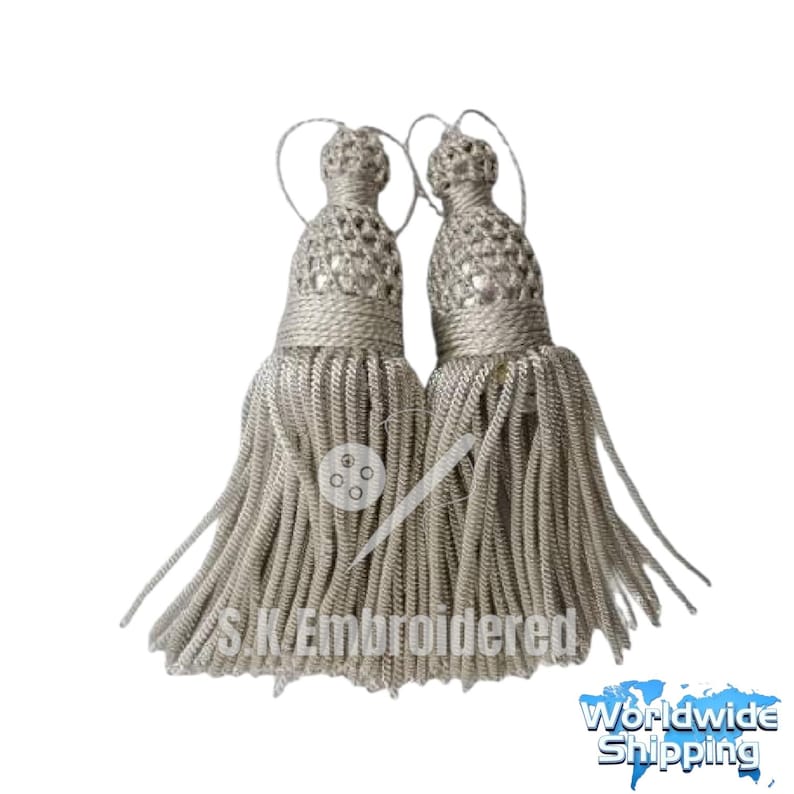 K&ouml;nnte beinhalten: Zwei silberfarbene Quasten mit aufwendig gewebten Oberteilen und langen, flie&szlig;enden Str&auml;hnen. Die Quasten sind durch eine d&uuml;nne, geschlungene Kordel verbunden. Der Text "S.K. Embroidered" ist auf der Vorderseite der Quasten zu sehen.