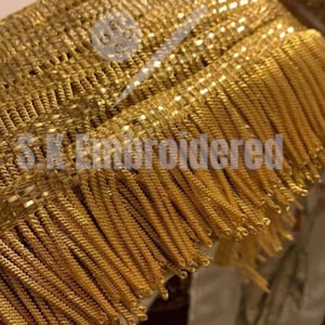 May include: Close-up of gold-coloured fringe trim with a woven base. The fringe is made of twisted strands, creating a textured appearance. The text "S.K. Embroidered" is visible on the trim.