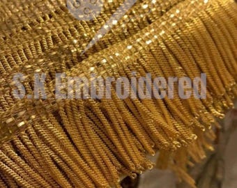 May include: Close-up of gold-coloured fringe trim with a woven base. The fringe is made of twisted strands, creating a textured appearance. The text "S.K. Embroidered" is visible on the trim.