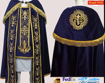 May include: A blue velvet cape with gold embroidery and fringe. The cape has a large gold embroidered design on the back with the letters "JHS" in the center. The cape is worn over a white garment.