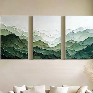 May include: Three framed abstract artworks showing a mountain range in shades of green and white. The art has a textured, three-dimensional look, with varying shades of green creating depth. The art is displayed above a white sofa in a living room.