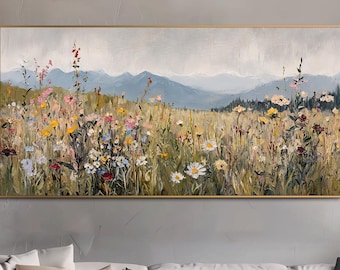 Op de afbeelding: Een ingelijst landschapsschilderij toont een veld met wilde bloemen in bloei, met bergen op de achtergrond. Het kunstwerk gebruikt groen, geel, roze en blauw, waardoor een serene scène ontstaat. Het schilderij wordt boven een witte bank weergegeven.