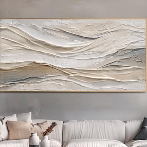 Puede incluir: Una pintura abstracta rectangular grande con marco dorado. La obra presenta un diseño texturizado y en capas en tonos blancos, beige y marrón, que recuerda a colinas o olas. Cuelga sobre un sofá blanco y una mesa de centro de madera.