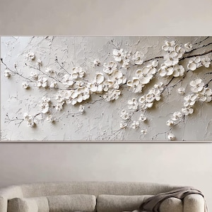 May include: A framed, textured artwork featuring white blossoms on dark branches against a light gray background. The blossoms are raised, creating a 3D effect. The artwork is rectangular and mounted on a wall above a beige sofa.