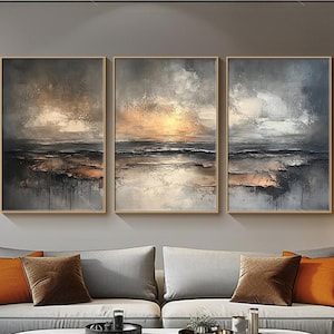 May include: Three abstract landscape paintings in gold, grey, and white tones. The paintings are framed in a thin, gold frame and are hung on a grey wall. The paintings depict a seascape with a sunset in the background.