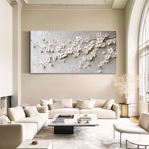 May include: A large, rectangular, textured artwork featuring a relief of white blossoms on dark branches against a gray background. The artwork is displayed above a white sofa in a modern living room setting.
