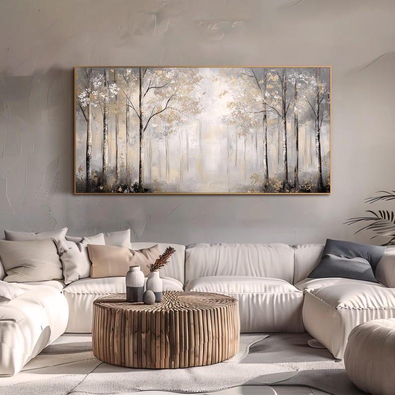 May include: A large abstract painting of a forest scene in shades of gray, white, and gold. The painting is framed in a thin gold frame and hangs on a gray wall above a white sectional sofa.