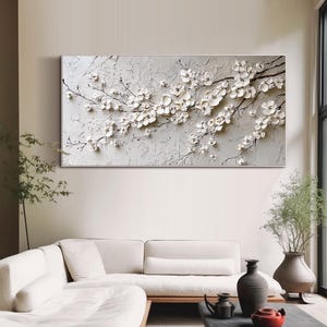 May include: A large rectangular canvas features a textured, light gray background with a relief sculpture of white cherry blossoms. The blossoms and branches extend across the canvas, creating a three-dimensional effect. The artwork is displayed above a white sofa.