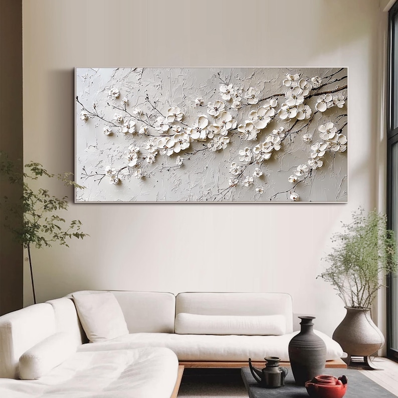 May include: A large rectangular canvas features a textured, light gray background with a relief sculpture of white cherry blossoms. The blossoms and branches extend across the canvas, creating a three-dimensional effect. The artwork is displayed above a white sofa.
