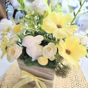 May include: A white ceramic bunny figurine sits in a wooden planter with yellow flowers, green moss, and yellow speckled Easter eggs. The planter is tied with a yellow ribbon.