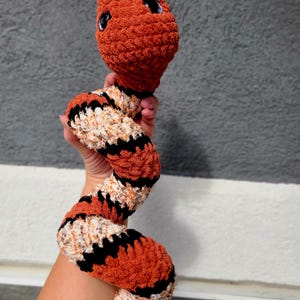 May include: A handmade orange, black, and cream-colored crochet snake toy with blue eyes. The snake is held up against a gray background. The snake's body is a mix of orange, black, and cream-colored yarn, creating a striped pattern.