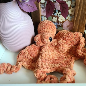 May include: A crochet octopus lovey made from soft, variegated orange and peach yarn. The plush toy features a rounded head, a flat, textured body, and curled tentacles. It has small, black, circular eyes stitched onto the head.
