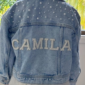 May include: Pale blue denim jacket adorned with pearls. The name "CAMILA" is spelled out on the back in pearl-covered letters. The jacket has a traditional design, including a collar, pockets, and button fastenings.