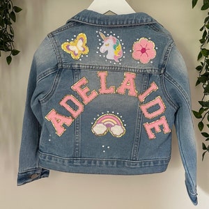May include: A blue denim jacket with pink and gold patches, including a unicorn, butterfly, flower, and rainbow. The name "ADELAIDE" is spelled out in pink letters on the back.
