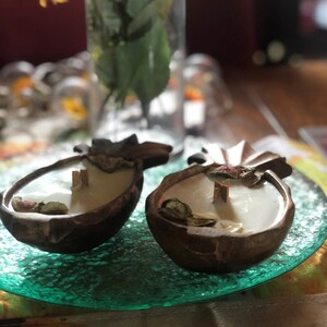 Op de afbeelding: Twee kokosnootschaal kaarsen met witte was en houten lonten staan op een groene glazen plaat. De bruine schelpen zijn versierd met gedroogde bloemen en bladeren. De achtergrond is wazig.