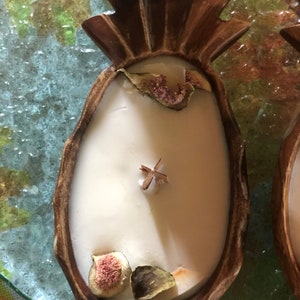 Op de afbeelding: Een ananasvormige kaarsenhouder van donkerbruin hout, gevuld met witte was. De kaars is versierd met gedroogde vijgen en een kaneelstokje. De kaars staat op een glazen oppervlak met een kleurrijke achtergrond.