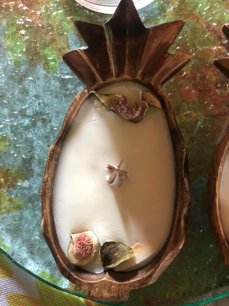 Op de afbeelding: Een ananasvormige kaarsenhouder van donkerbruin hout, gevuld met witte was. De kaars is versierd met gedroogde vijgen en een kaneelstokje. De kaars staat op een glazen oppervlak met een kleurrijke achtergrond.