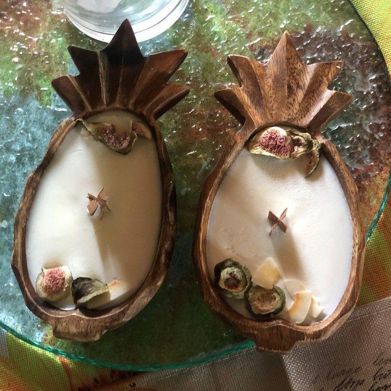 Op de afbeelding: Twee kaarsen in ananasvorm met houten toppen. De kaarsen zijn gevuld met witte was en versierd met gedroogd fruit. De houten bases zijn bruin van kleur. De kaarsen staan op een glazen oppervlak.