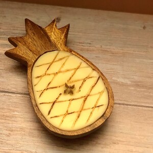 Op de afbeelding: Een houten ananasvormige kandelaar met een lichtgele kaars erin. De kaars heeft een kruispatroon en een klein stervormig accent. De houder is gemaakt van lichtbruin hout en staat op een houten oppervlak.