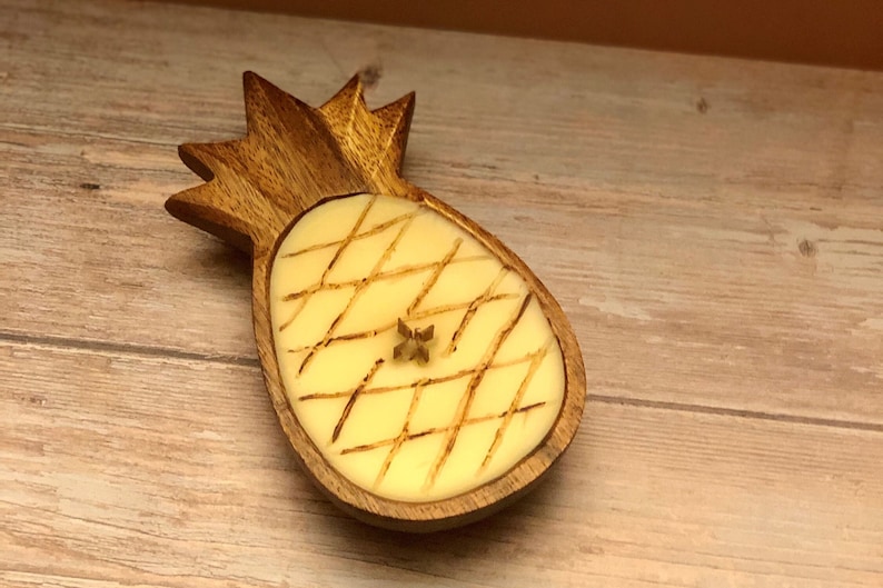 Op de afbeelding: Een houten ananasvormige kandelaar met een lichtgele kaars erin. De kaars heeft een kruispatroon en een klein stervormig accent. De houder is gemaakt van lichtbruin hout en staat op een houten oppervlak.