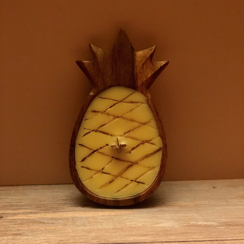 Op de afbeelding: Een ananasvormige kaarsenhouder van donker hout, met een gele kaars erin. De kaars heeft een kruisvormige lont en een ruitpatroon. De kaarsenhouder staat op een houten oppervlak.