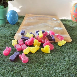 May include: An assortment of colorful, bunny-shaped candies in shades of pink, yellow, and purple are scattered on a bed of green artificial grass. A clear cellophane bag is partially open, with more candies spilling out. Easter-themed.