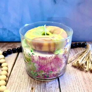 May include: A clear glass jar candle with a green and pink donut-shaped candle inside. The donut candle has a white wick and is surrounded by pink and green glitter.