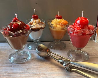 May include: Four decorative ice cream sundae candles in clear glass dishes. Each candle has a different color and topping, including chocolate, caramel, and cherries. A silver ice cream scoop rests in front of the candles.