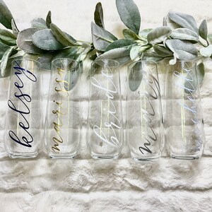 Op de afbeelding: Vijf heldere glazen champagneflutes met kalligrafie-lettering. De flutes zijn in een rij op een wit, pluche oppervlak gerangschikt. De letters op de flutes luiden "Kelsey", "marissa", "bride", "mrs" en "miss hello".