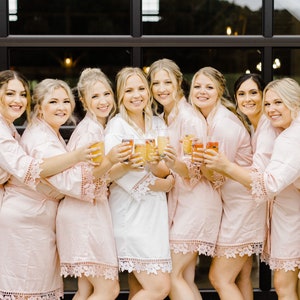 Op de afbeelding: Een groep van acht vrouwen die roze satijnen gewaden met kant dragen, champagne glazen omhoog houden en naar de camera lachen.
