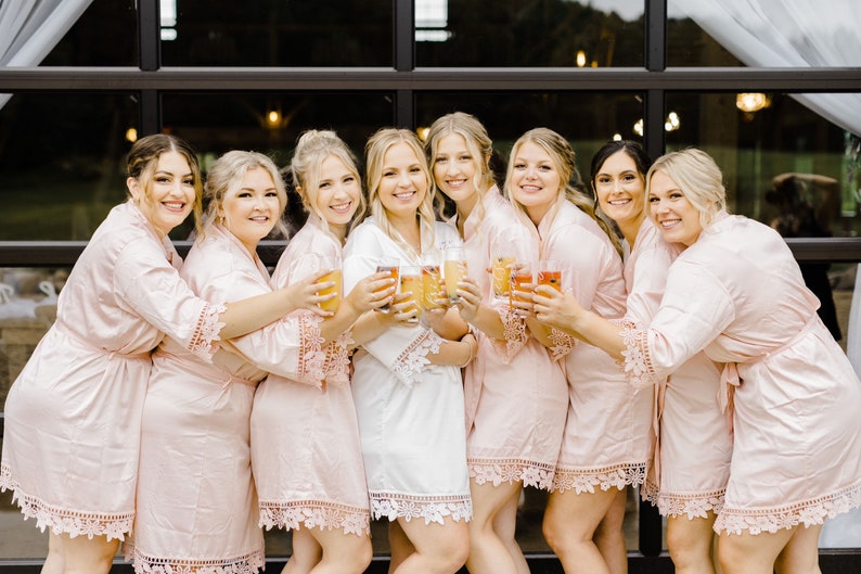Op de afbeelding: Een groep van acht vrouwen die roze satijnen gewaden met kant dragen, champagne glazen omhoog houden en naar de camera lachen.