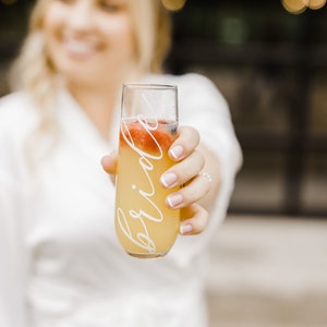 Op de afbeelding: Een heldere glazen champagnefluit met het woord "bride" erop. De fluit is gevuld met een gele vloeistof en heeft rood fruit erin.