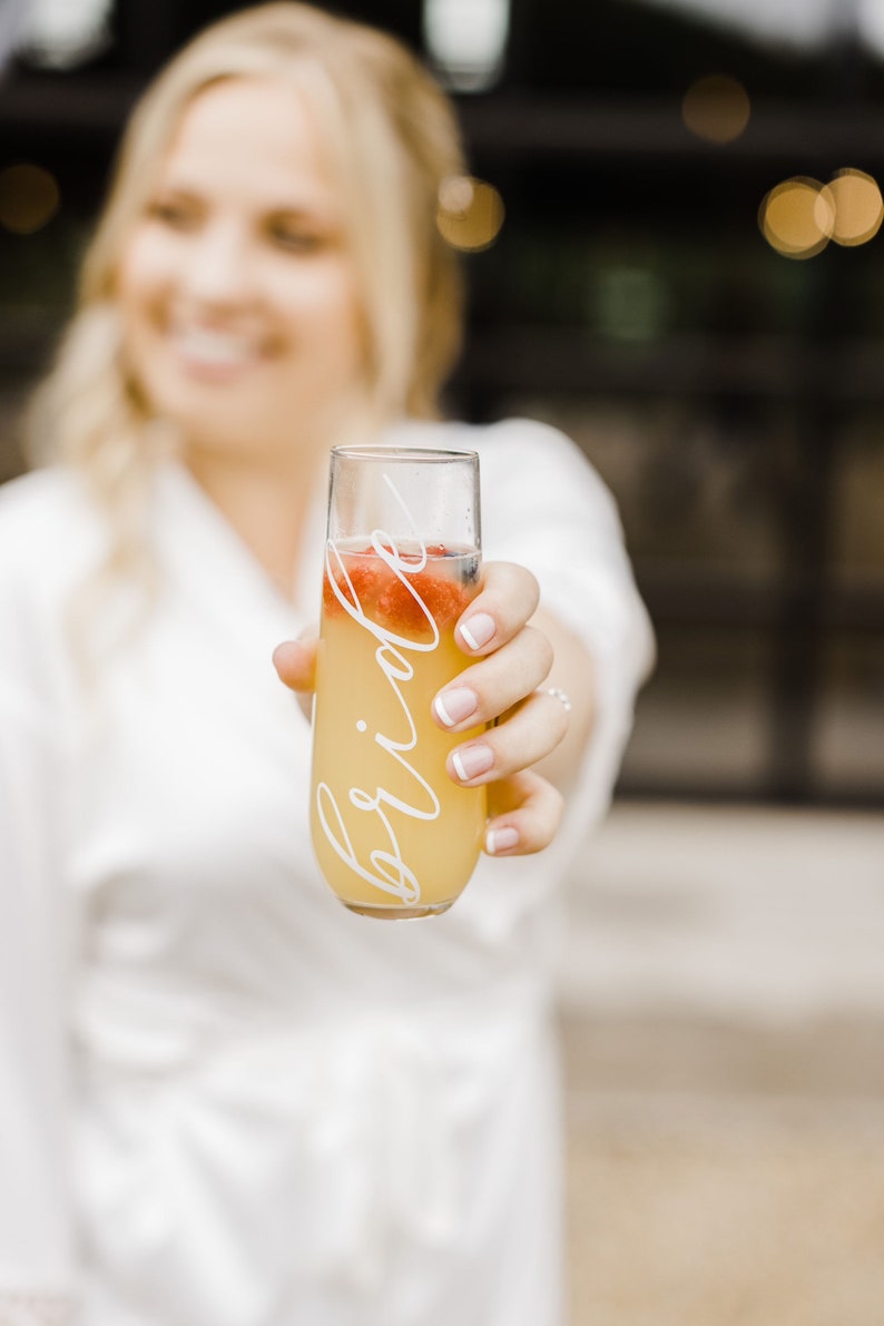 Op de afbeelding: Een heldere glazen champagnefluit met het woord "bride" erop. De fluit is gevuld met een gele vloeistof en heeft rood fruit erin.