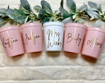 May include: Five pink and white plastic cups with gold lettering. The cups are labeled "Kaitlynn", "Jaclynn", "Mrs. Weeks", "Bailey", and "Alexis".