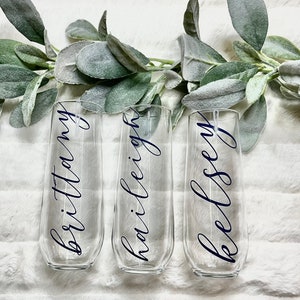 Op de afbeelding: Drie heldere glazen champagneflutes met de namen "Brittany", "Hailey" en "Kelsey" geschreven in zwarte kalligrafie.