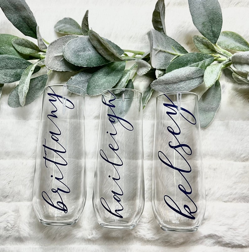 Op de afbeelding: Drie heldere glazen champagneflutes met de namen "Brittany", "Hailey" en "Kelsey" geschreven in zwarte kalligrafie.