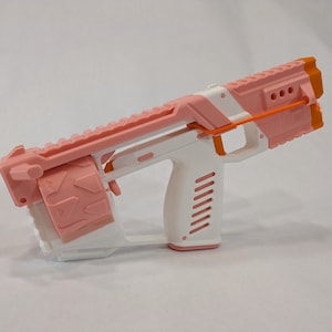 May include: A pink and white toy gun with an orange strap. The gun has a cylindrical magazine and a textured grip.