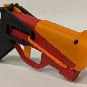 May include: A red and orange toy gun with a black grip and a textured surface. The gun has a strap attached to it and the word "TESTME" is printed on the side.