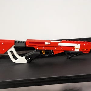 May include: A red and white toy gun with a black grip and orange barrel. The gun has a white stripe on the top and a black trigger.