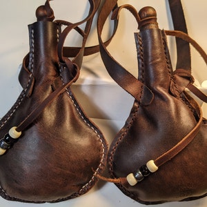 May include: Two brown leather water pouches with attached straps. The pouches have a teardrop shape and are hand-stitched with white thread. Each pouch has a leather strap with decorative beads.
