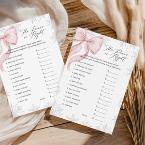 May include: Two baby shower game cards with the text "The Price is Right" and a pink bow illustration. The cards have a list of baby items with blank spaces for prices. The background is a wooden surface with dried plants.