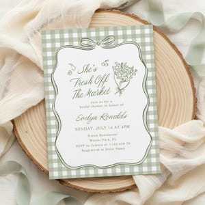 May include: A rectangular bridal shower invitation card with a sage green and white gingham border. The center features a white scalloped frame with a bow at the top, floral illustrations, and text reading: She's Fresh Off The Market, Evelyn Ronalds.