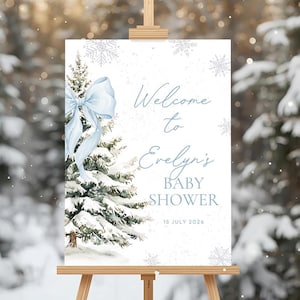 May include: A baby shower welcome sign on a wooden easel. The sign features a watercolor illustration of a snow-covered evergreen tree with a light blue bow. Text reads "Welcome to Evelyn's BABY SHOWER" and the date.
