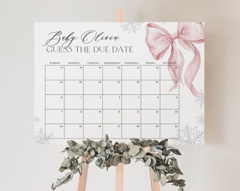 Op de afbeelding: Een witte kalender met de tekst "Baby Olivia Guess the Due Date" en een roze strik illustratie. Het kalenderraster is gevuld met data en sneeuwvlokontwerpen bevinden zich in de hoeken. De kalender wordt weergegeven op een houten ezel.